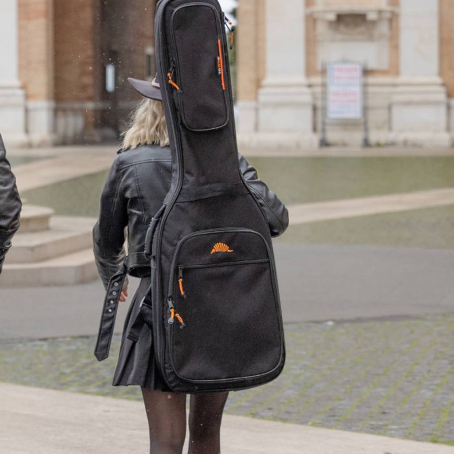 A bass player carrying their electric bass in a TOBAGO soft case, heading to perform at a concert in a square.